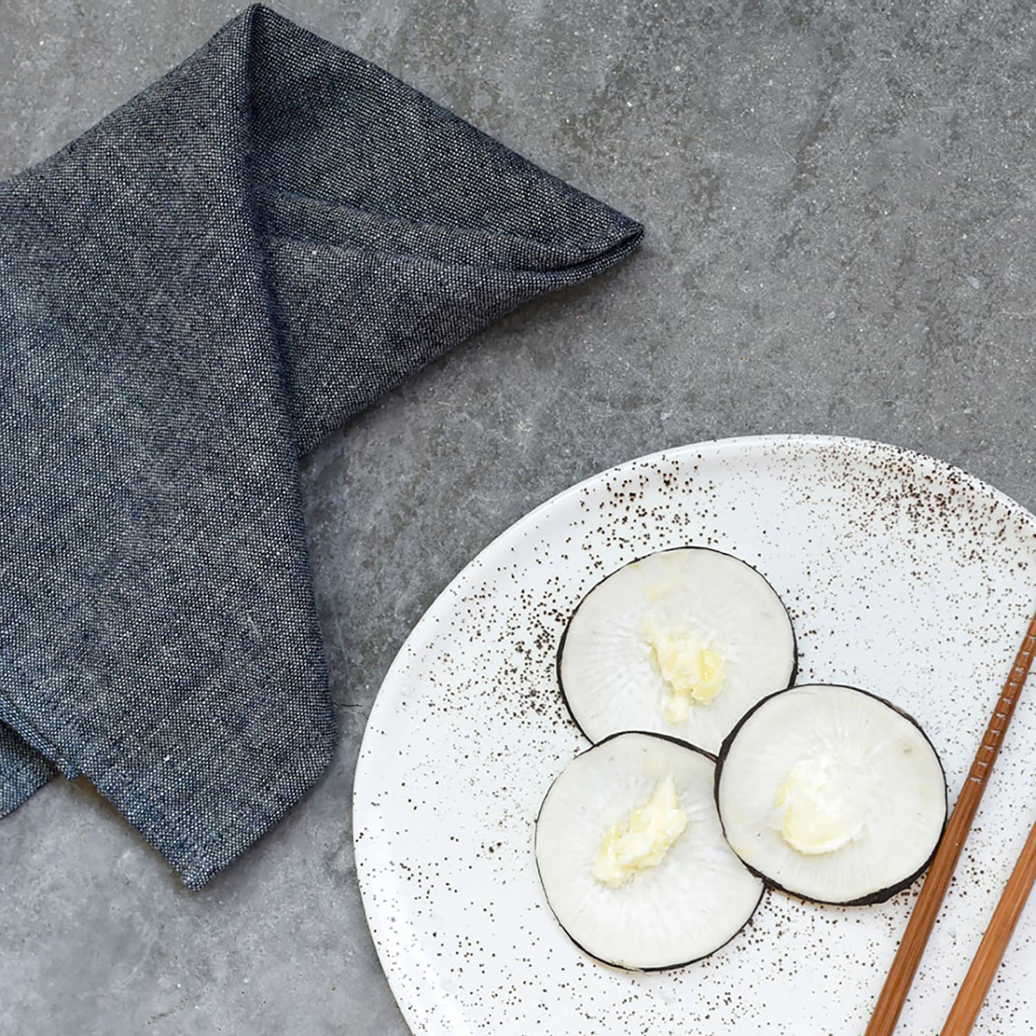 Three slices of radish on a speckled ceramic plate with chopsticks and a ATELIER SAUCIER chambray napkin on a cement surface.
